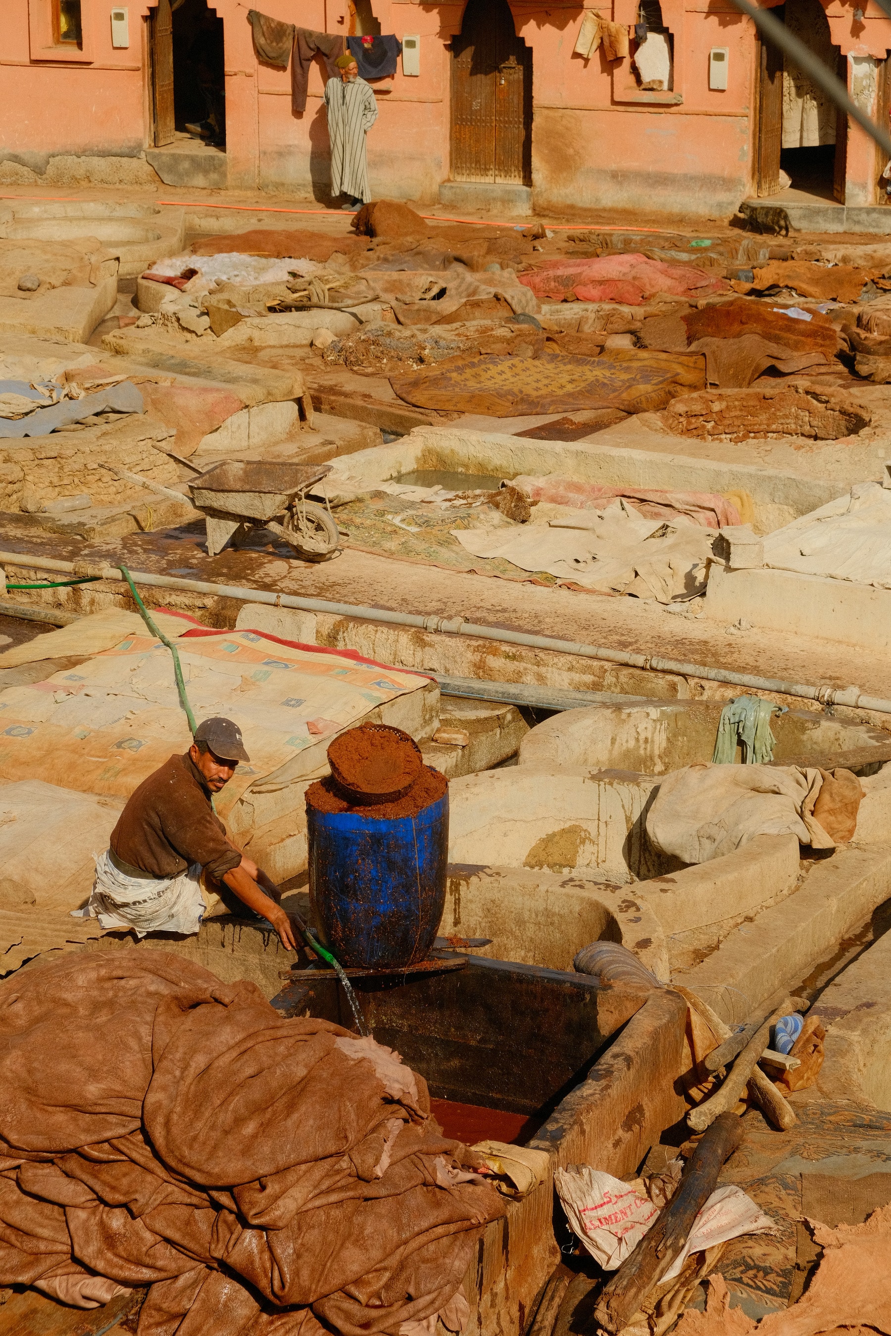 A tanner draws water beside a blue barrel at the pits of the Marrakech tanneries, with hides heaped around him