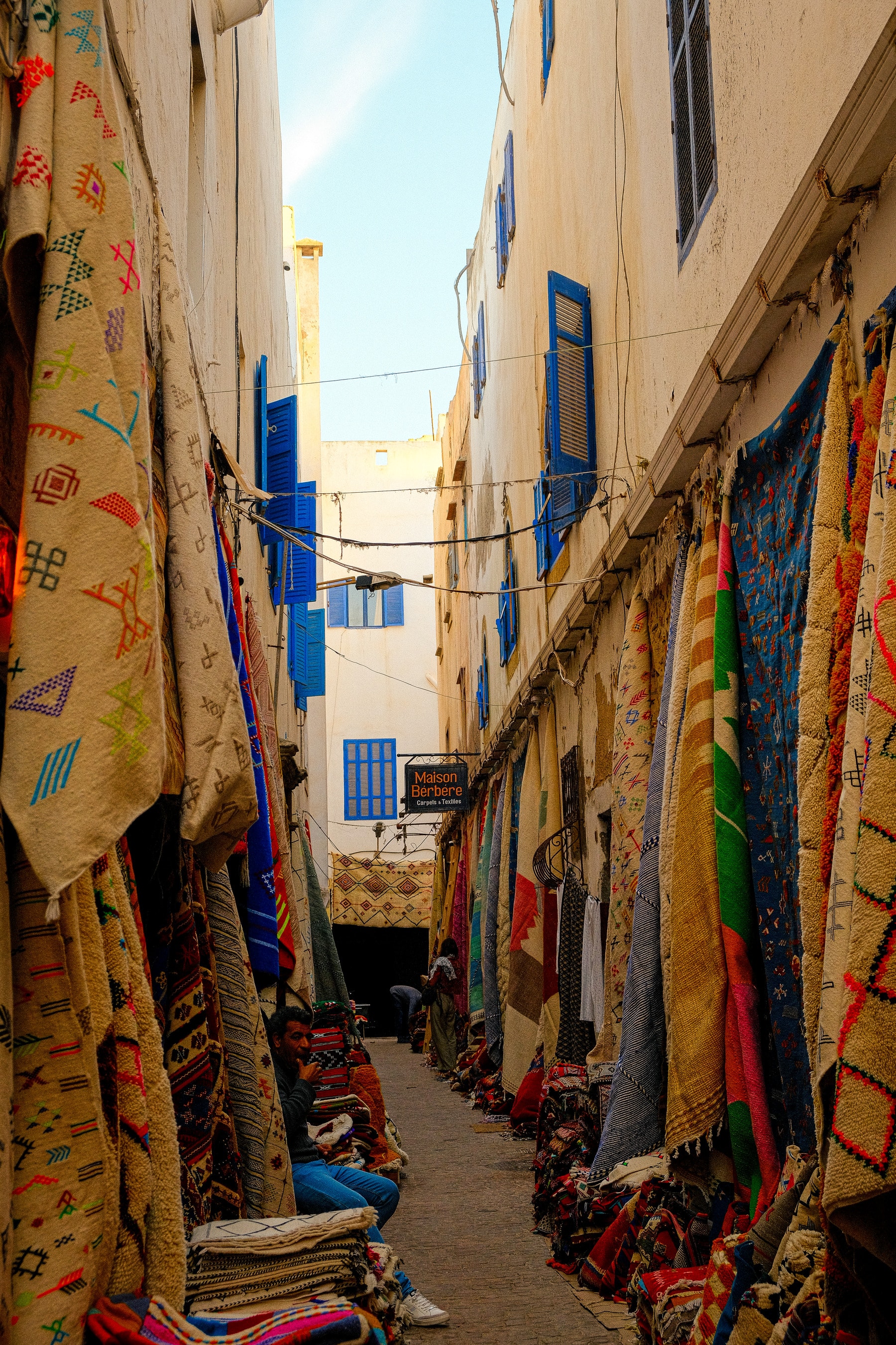 Rugs and blankets hang from ground to rooftop along a narrow alley of the Essaouira medina, between whitewashed walls and blue shutters