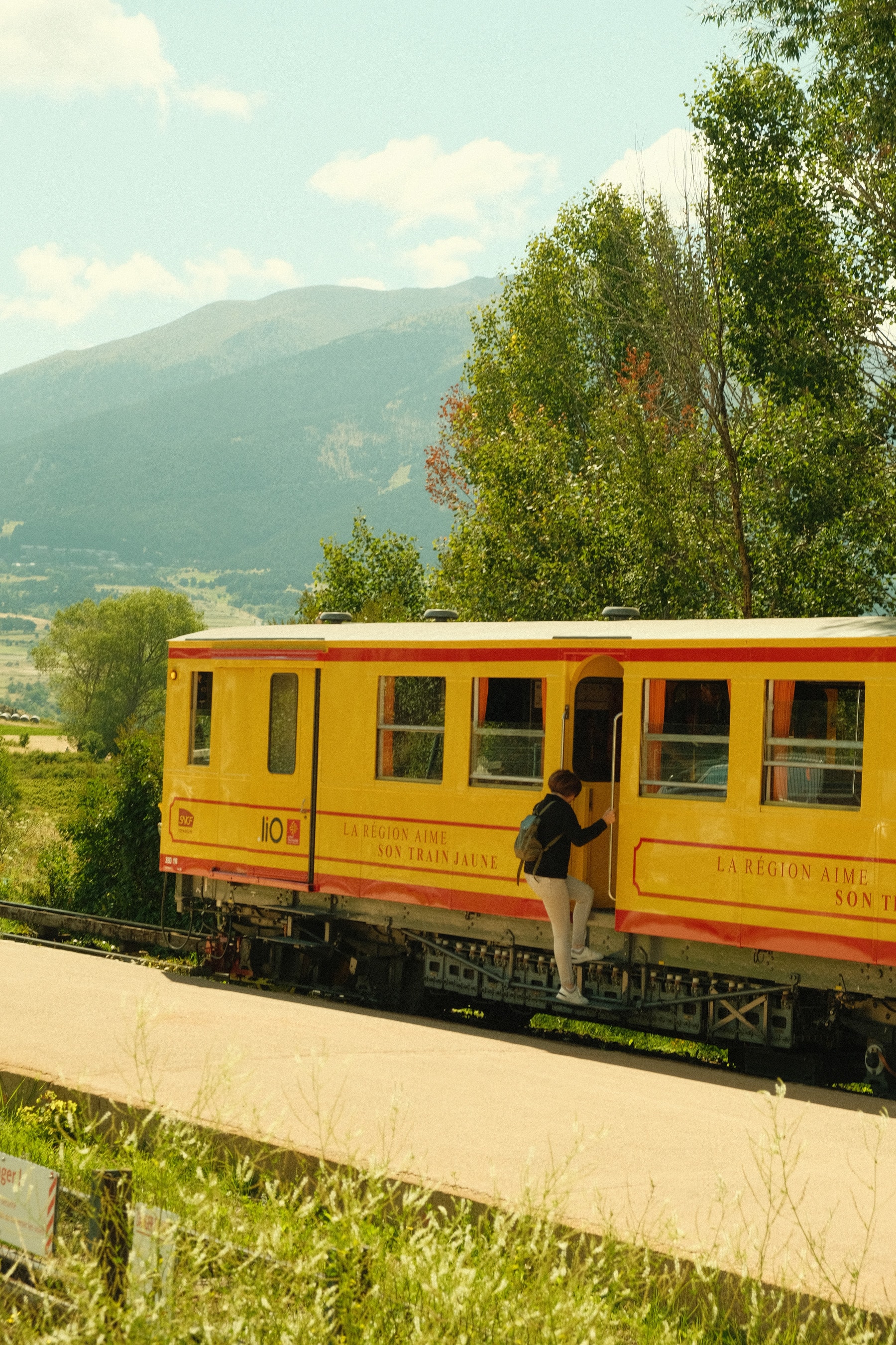 A passenger climbs aboard the Train Jaune through its open door, the yellow railcar small against the trees and the Pyrenees