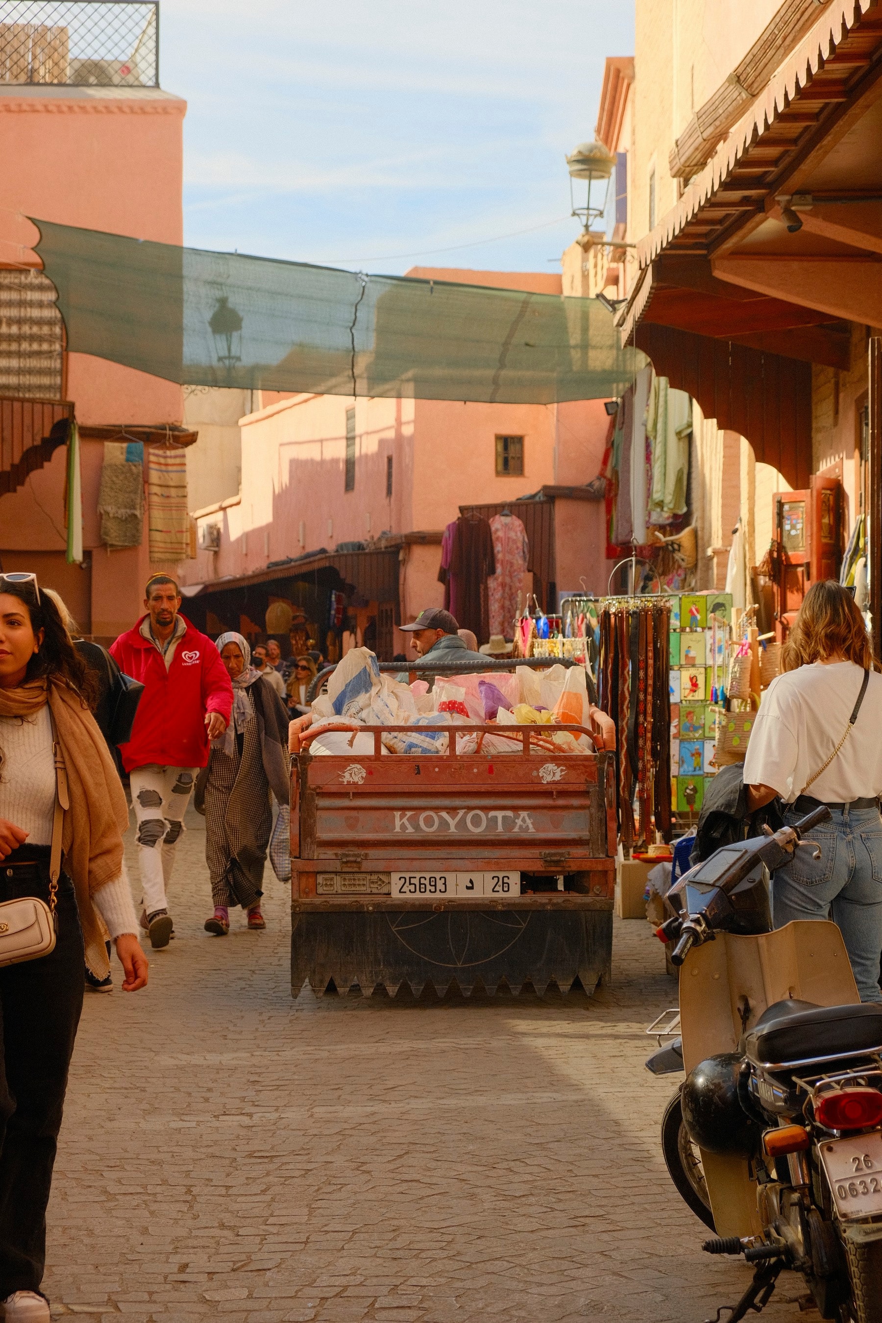 A busy souk lane in the Marrakech medina: a loaded motor-cart pushes through shoppers beneath awnings and a canvas shade, pink walls rising behind