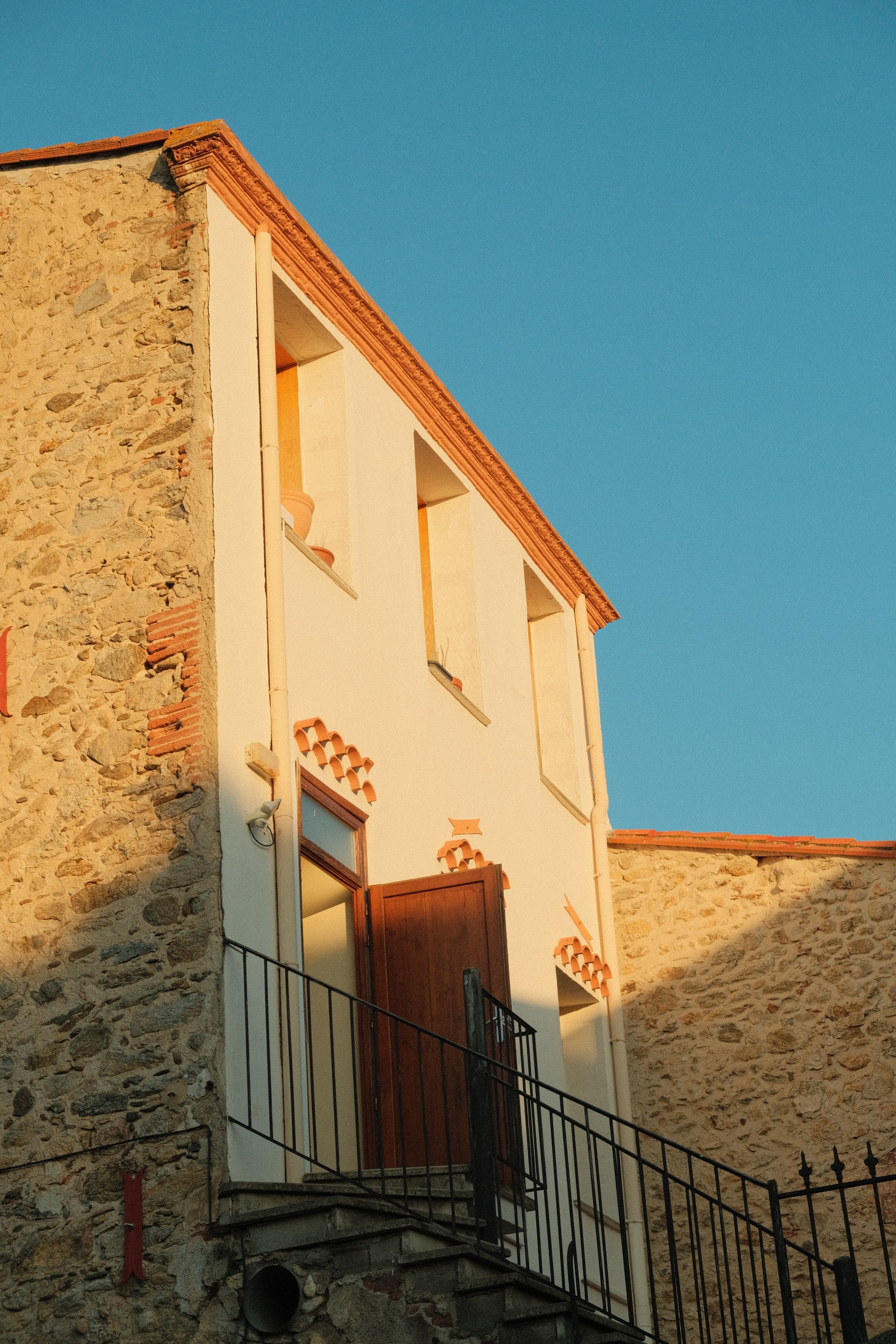 A stone-and-stucco house in Laroque-des-Albères in the last light of the day, a red door at the top of an iron stair and terracotta tiles set into the wall
